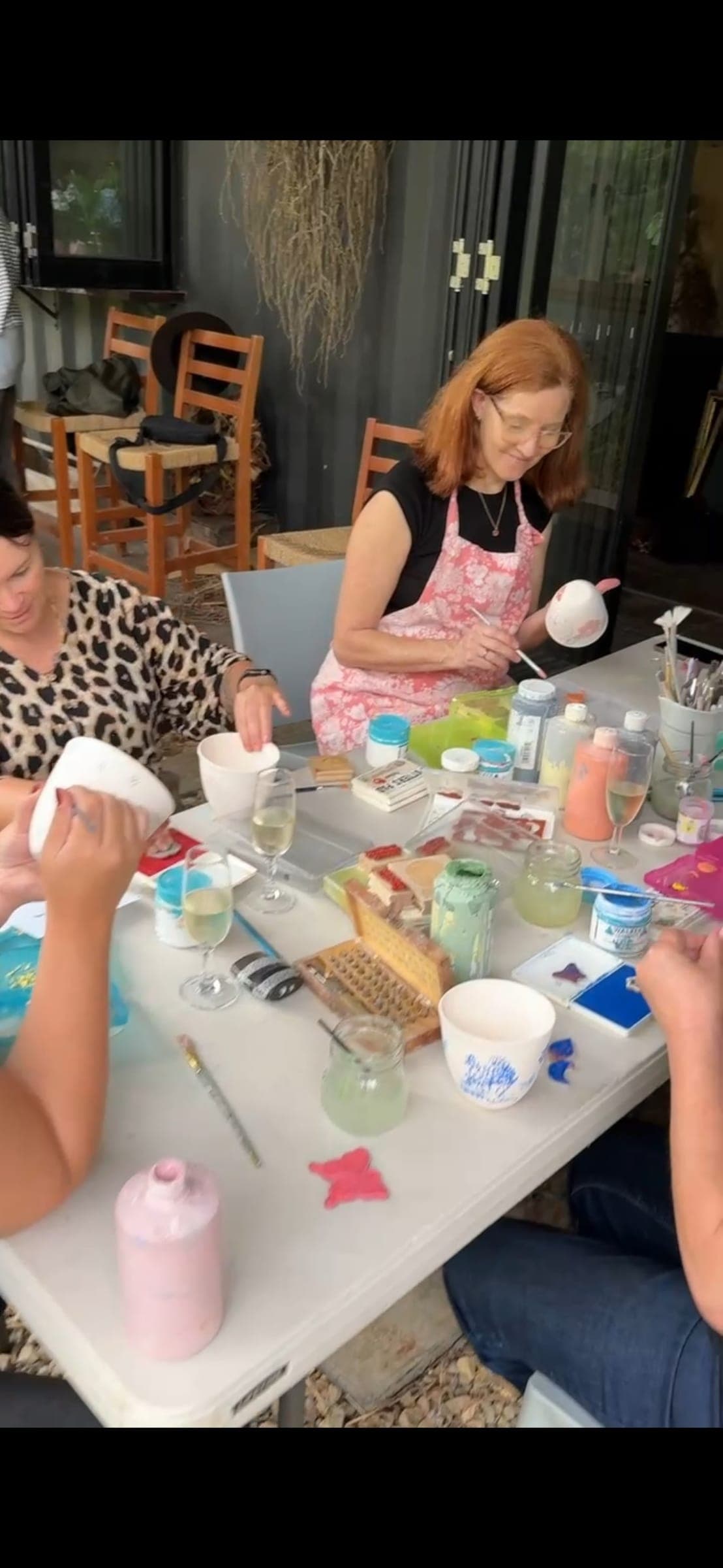A table of people painting cups and bowls, with wine glasses and paint pots between them