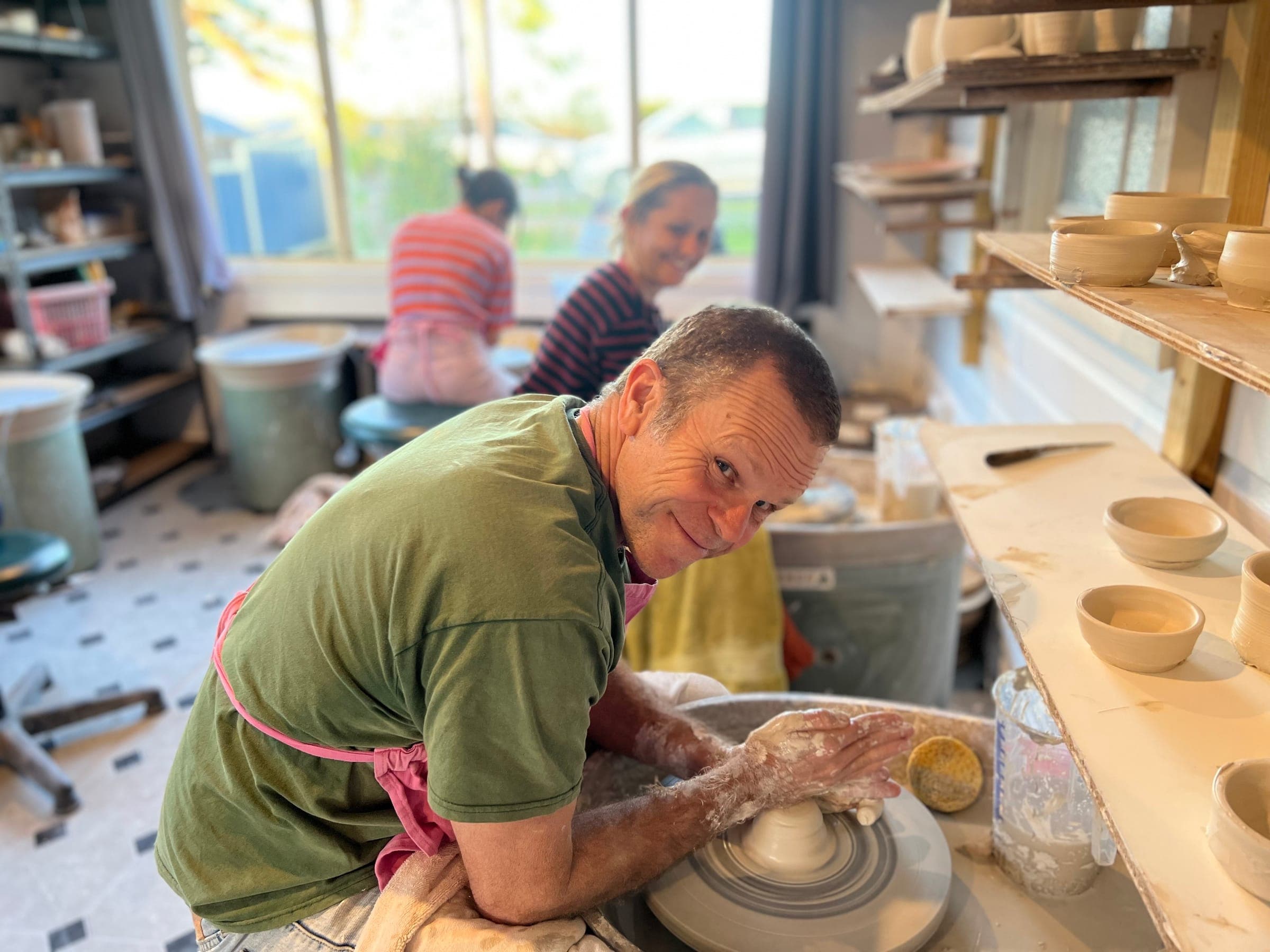 A potter smiling at the camera while shaping clay on the wheel
